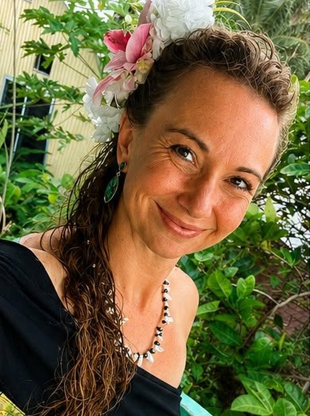 Portrait of Kelley Tagarino smiling, wearing flowers in her hair, with tropical greenery behind her.